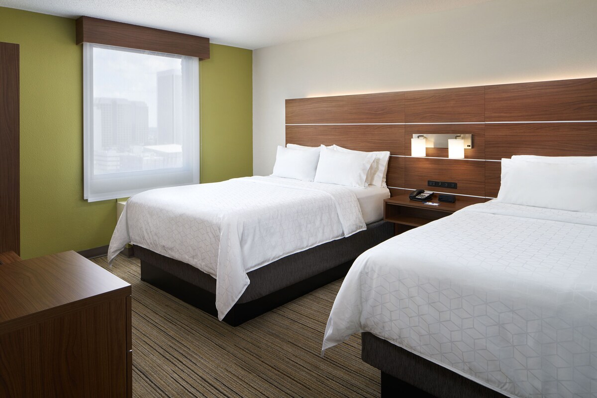 A hotel room with two beds, a nightstand, a telephone, and a window with a view of buildings.