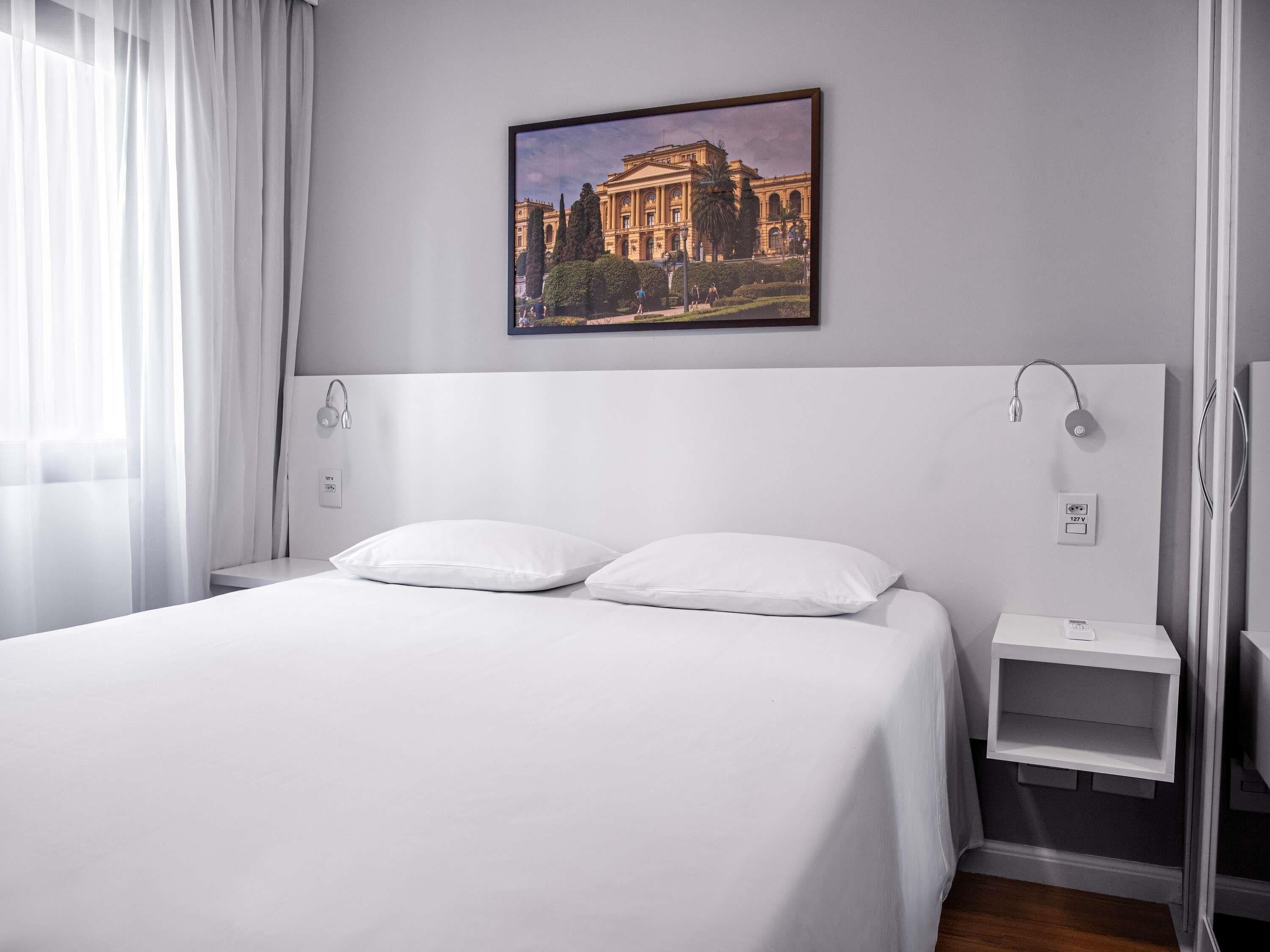 Une chambre d’hôtel avec un grand lit, une table de chevet, une lampe fixée au mur, une prise de courant et un tableau représentant un bâtiment.