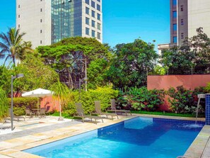Una piscina con sillas de descanso, rodeada de vegetación y edificios altos.