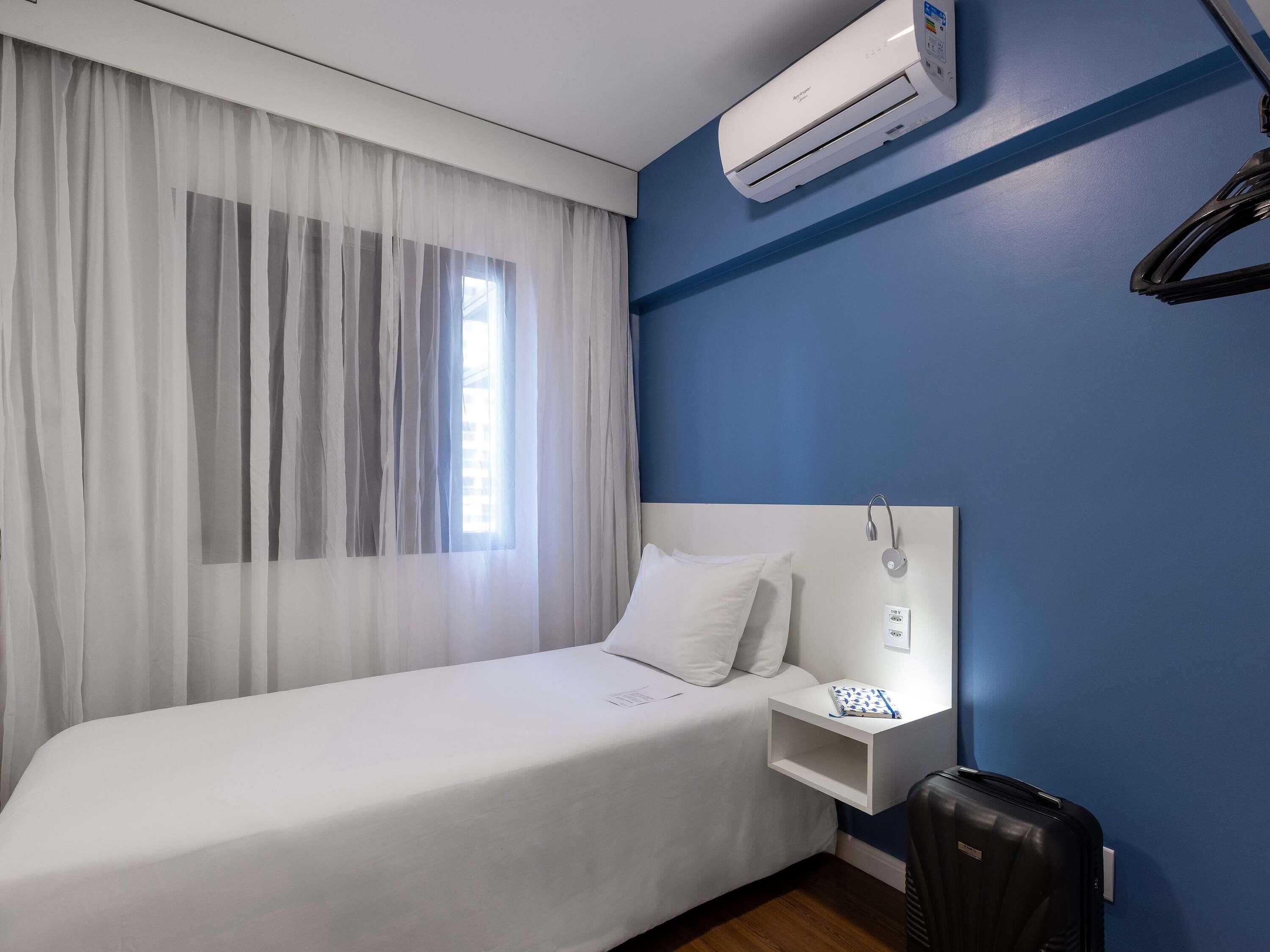 A hotel room with a bed, a bedside table, an air conditioning unit, a window with curtains, and a wall-mounted telephone.