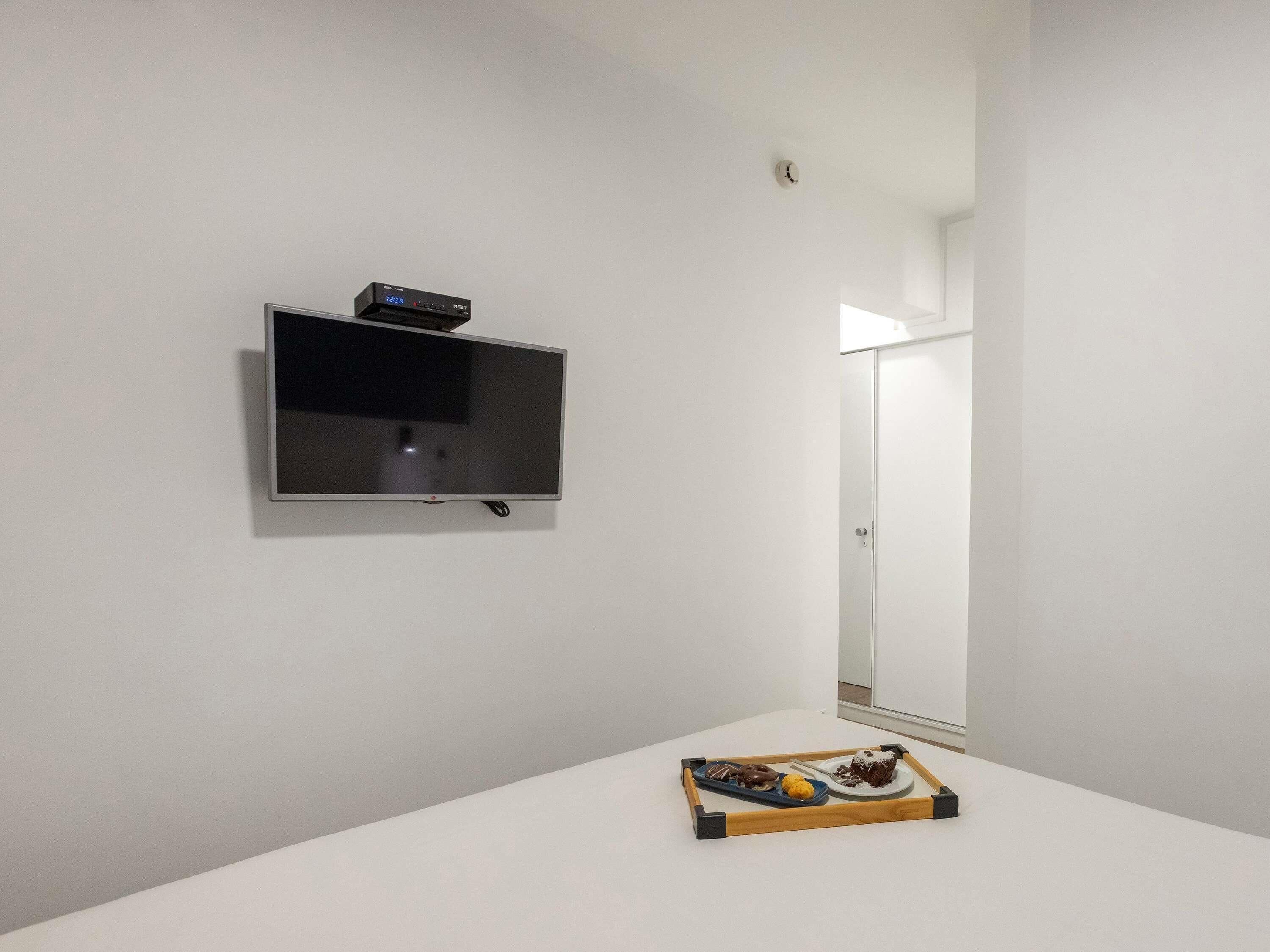 Een minimalistisch ingerichte kamer met een flatscreen televisie aan de muur, een klein houten dienblad met gebak op een witte tafel, en een deur die naar een andere kamer leidt.