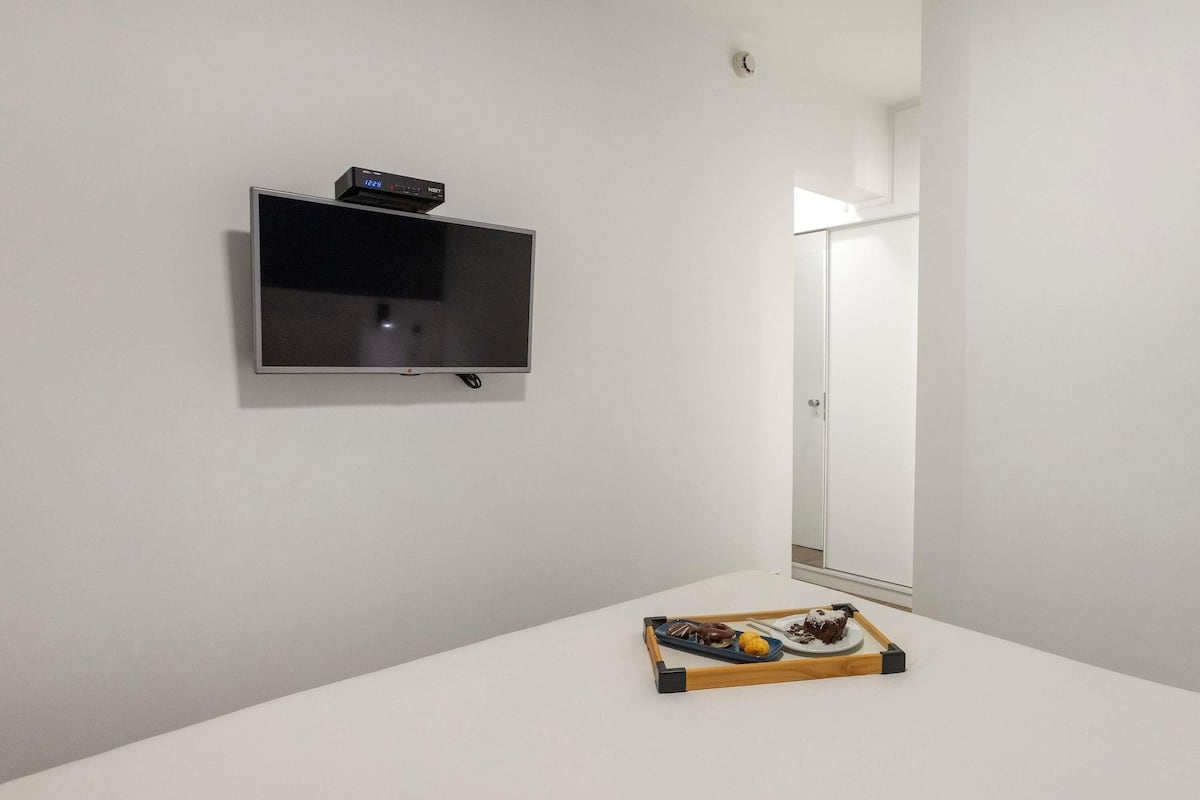 A minimalist room with a flat-screen TV mounted on the wall, a small wooden tray with pastries on a white table, and a door leading to another room.