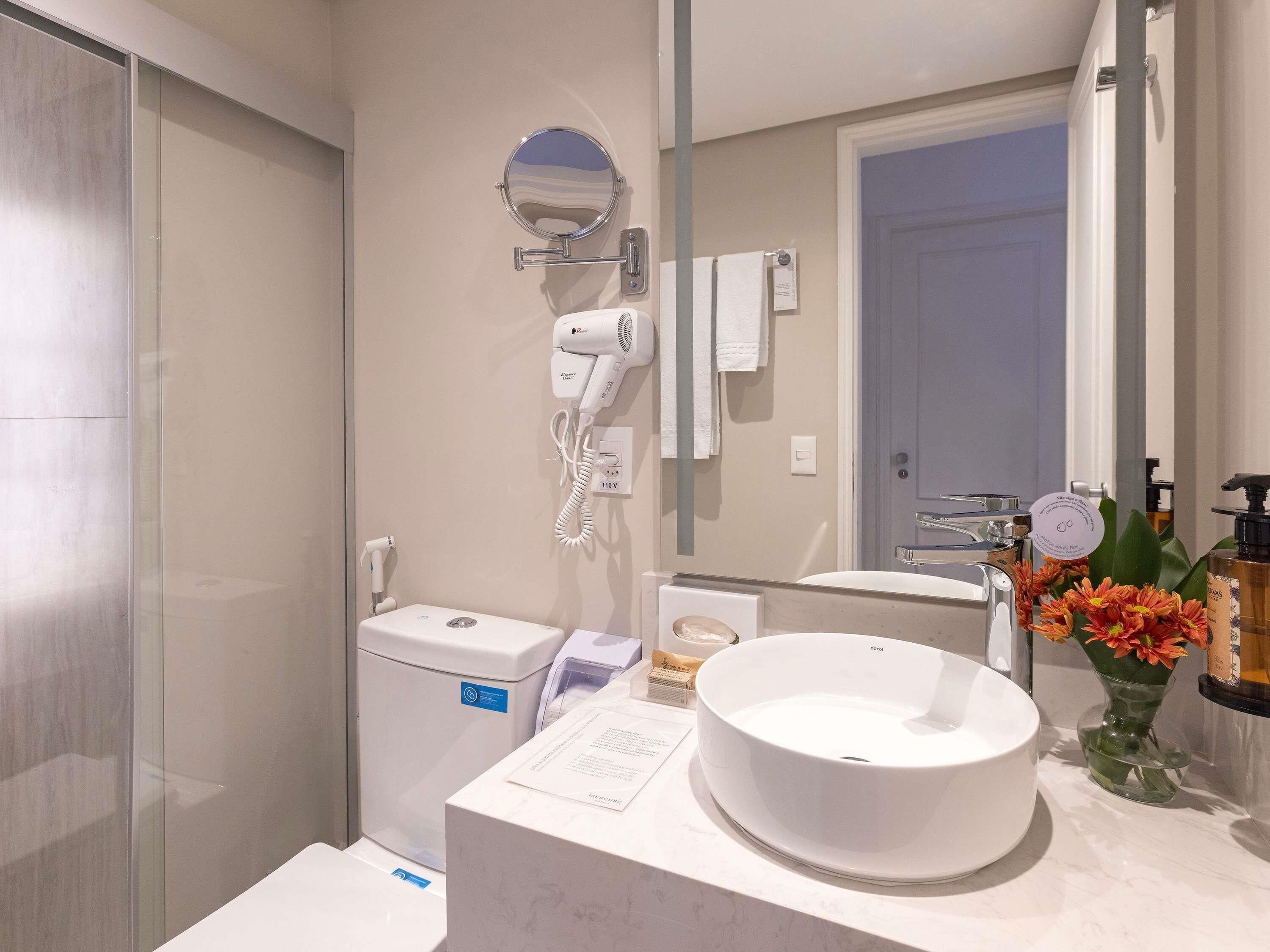 Een moderne badkamer met toilet, wastafel, spiegel, haardroger en een vaas met bloemen.