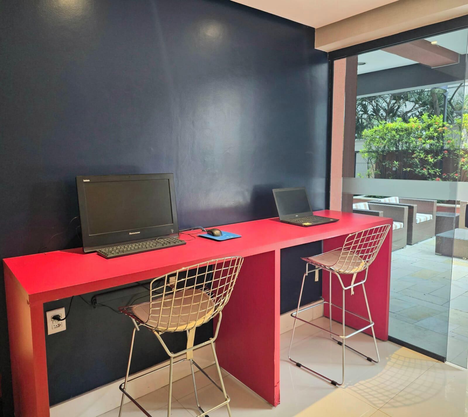 Un espacio de trabajo moderno con dos computadoras portátiles sobre un escritorio rojo, dos sillas de estructura metálica y un amplio ventanal que ofrece vistas al verdor exterior.