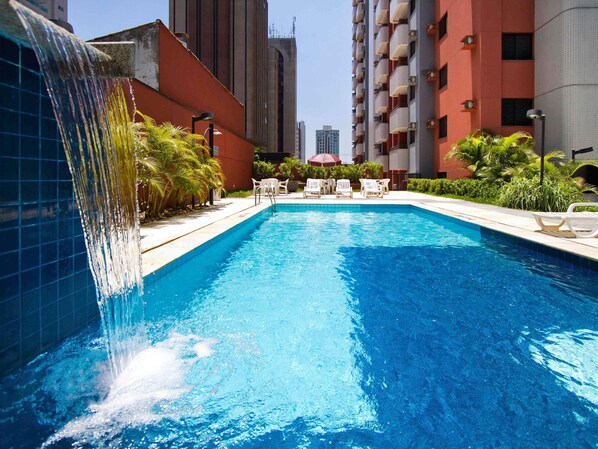 A swimming pool with a waterfall feature, surrounded by lounge chairs and greenery, with high-rise buildings in the background.