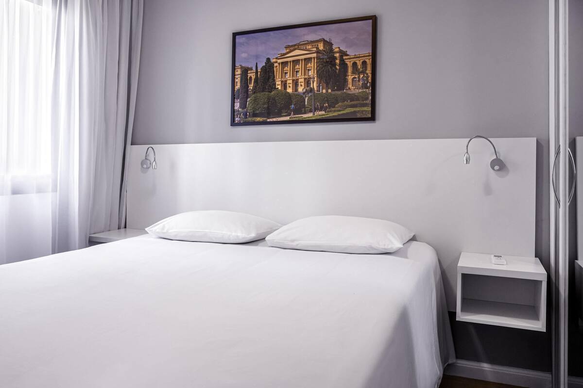 Una camera d'hotel con un letto grande, un comodino, un quadro appeso al muro e una finestra con tende.