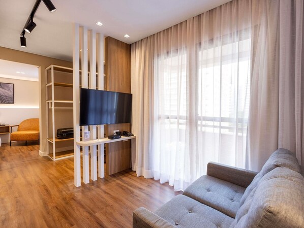 Un salon moderne doté d’un téléviseur à écran plat, d’un canapé, d’un parquet et d’une grande fenêtre ornée de rideaux légers.
