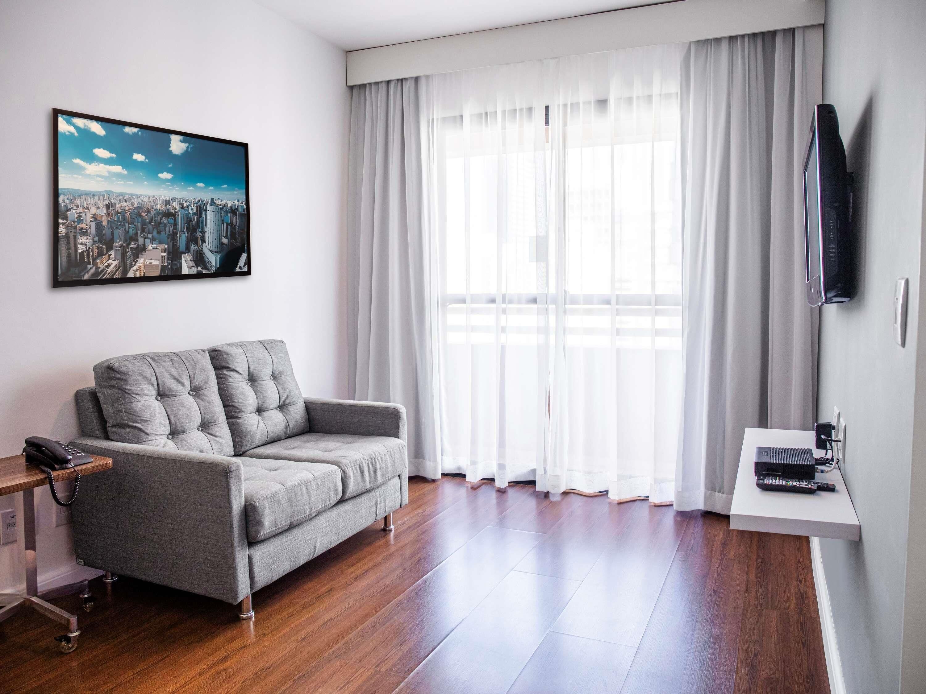 Een moderne woonkamer met een grijze bank, een stadslandschap schilderij, een groot raam met witte gordijnen en een wandgemonteerde televisie.