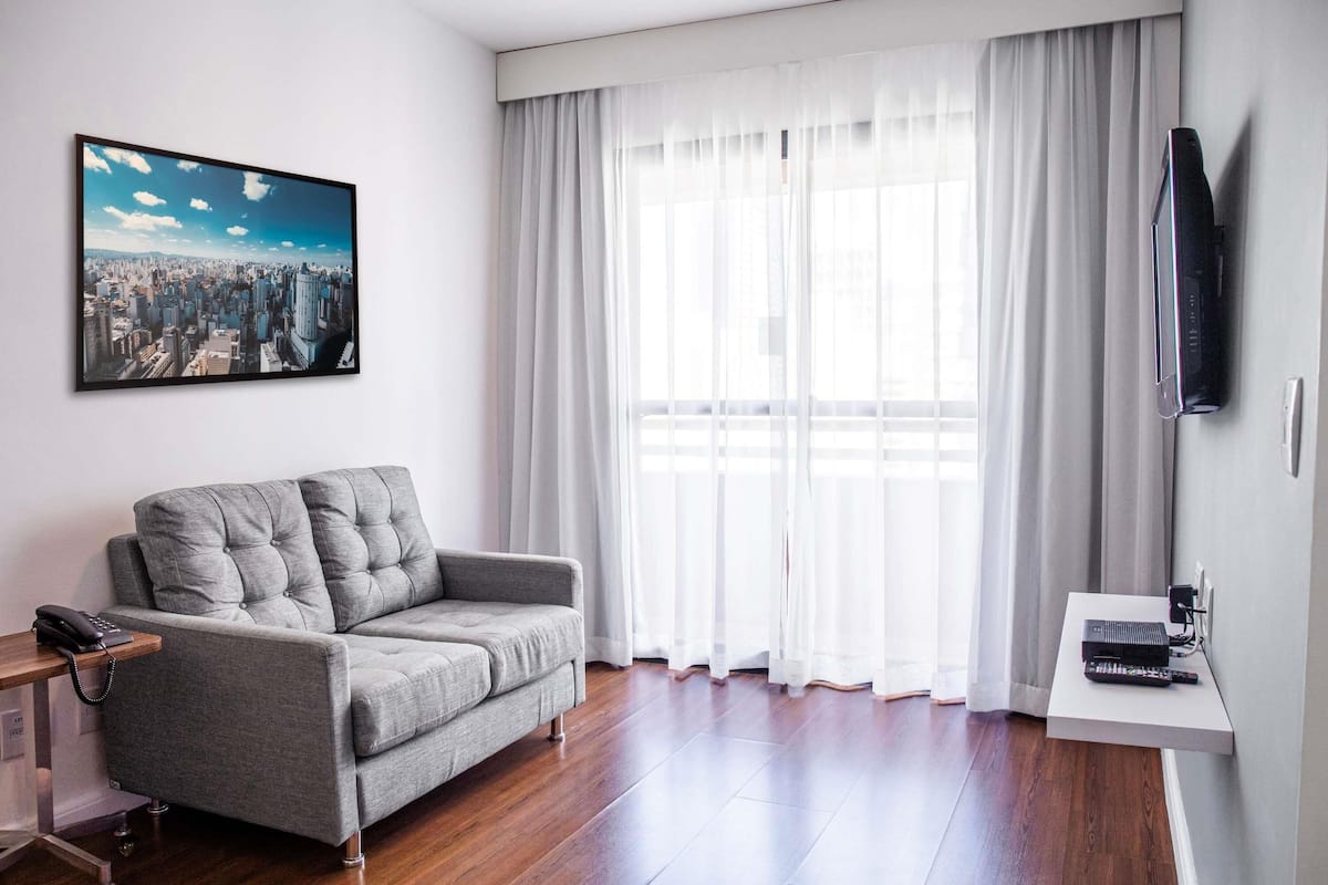 A modern living room with a grey sofa, a cityscape painting, a large window with white curtains, and a mounted television.