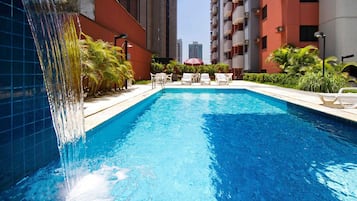 A swimming pool with a waterfall feature, surrounded by lounge chairs and greenery, with tall buildings in the background.