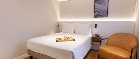 A hotel room with a bed, bedside table, chair, and an air conditioning unit.