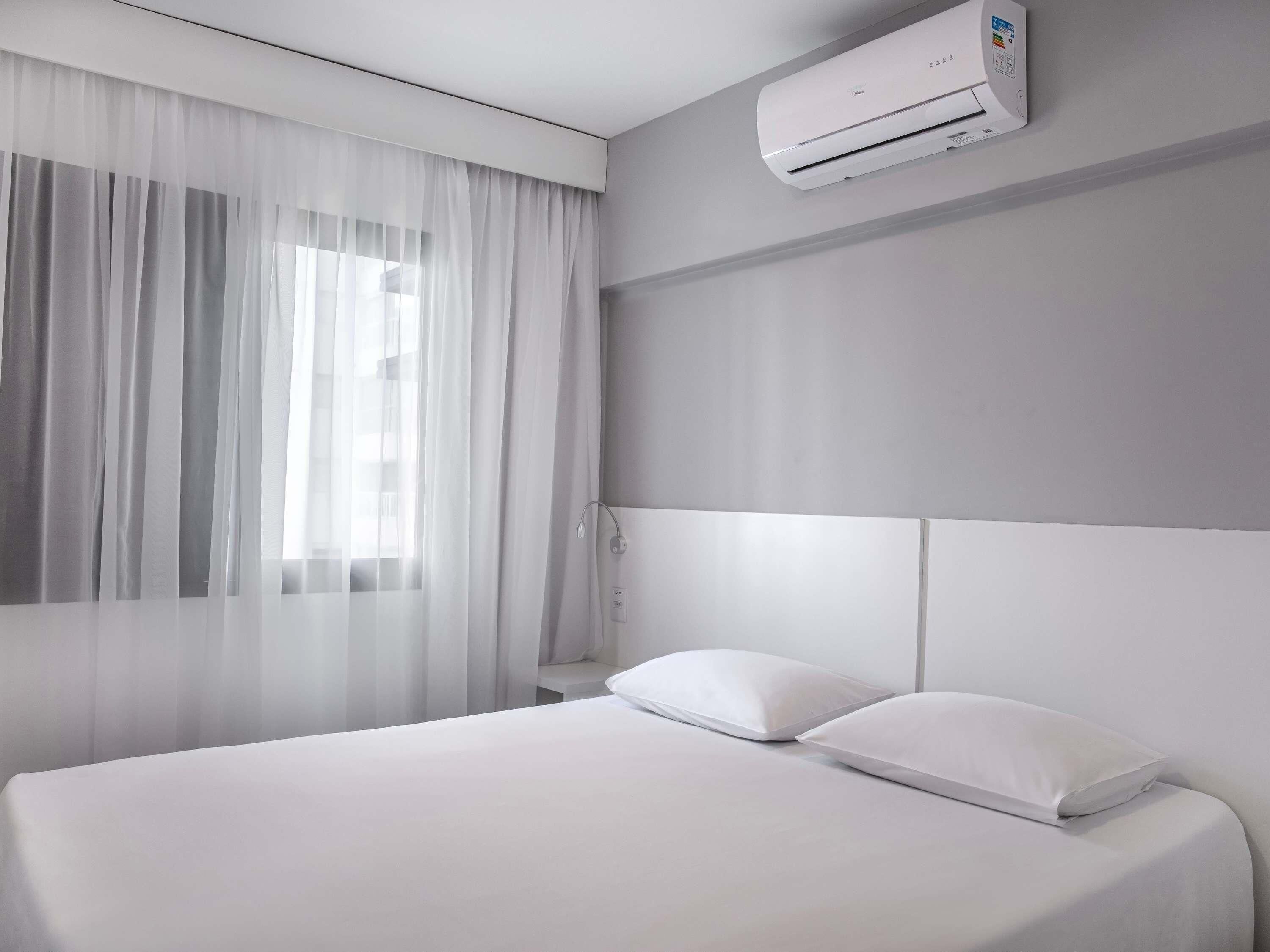 Une chambre d’hôtel avec un lit, des rideaux blancs, un appareil de climatisation et une table de chevet.