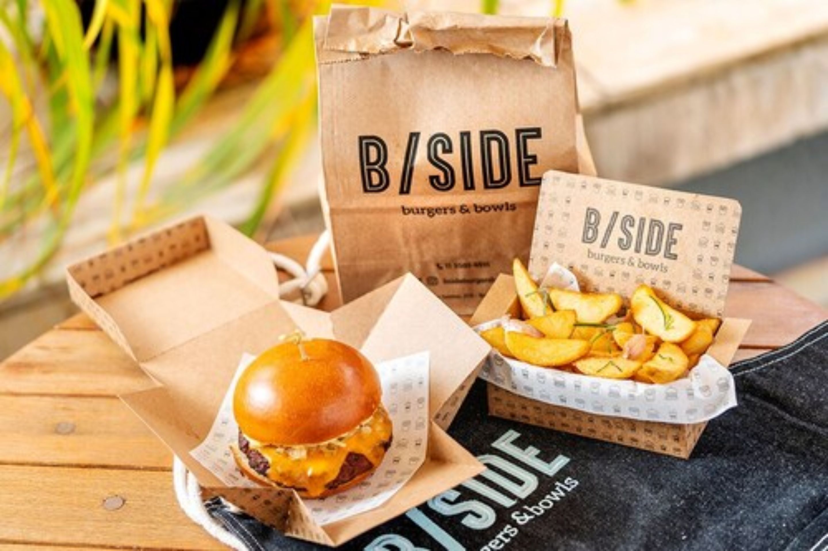 A meal from B/SIDE burgers & bowls, including a burger and fries, on a wooden table.