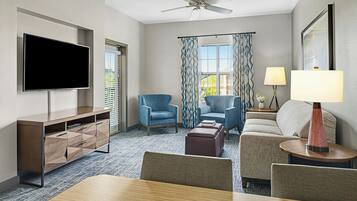 A modern hotel room with a flat-screen TV, a ceiling fan, a sofa, two armchairs, a coffee table, and a dining table.