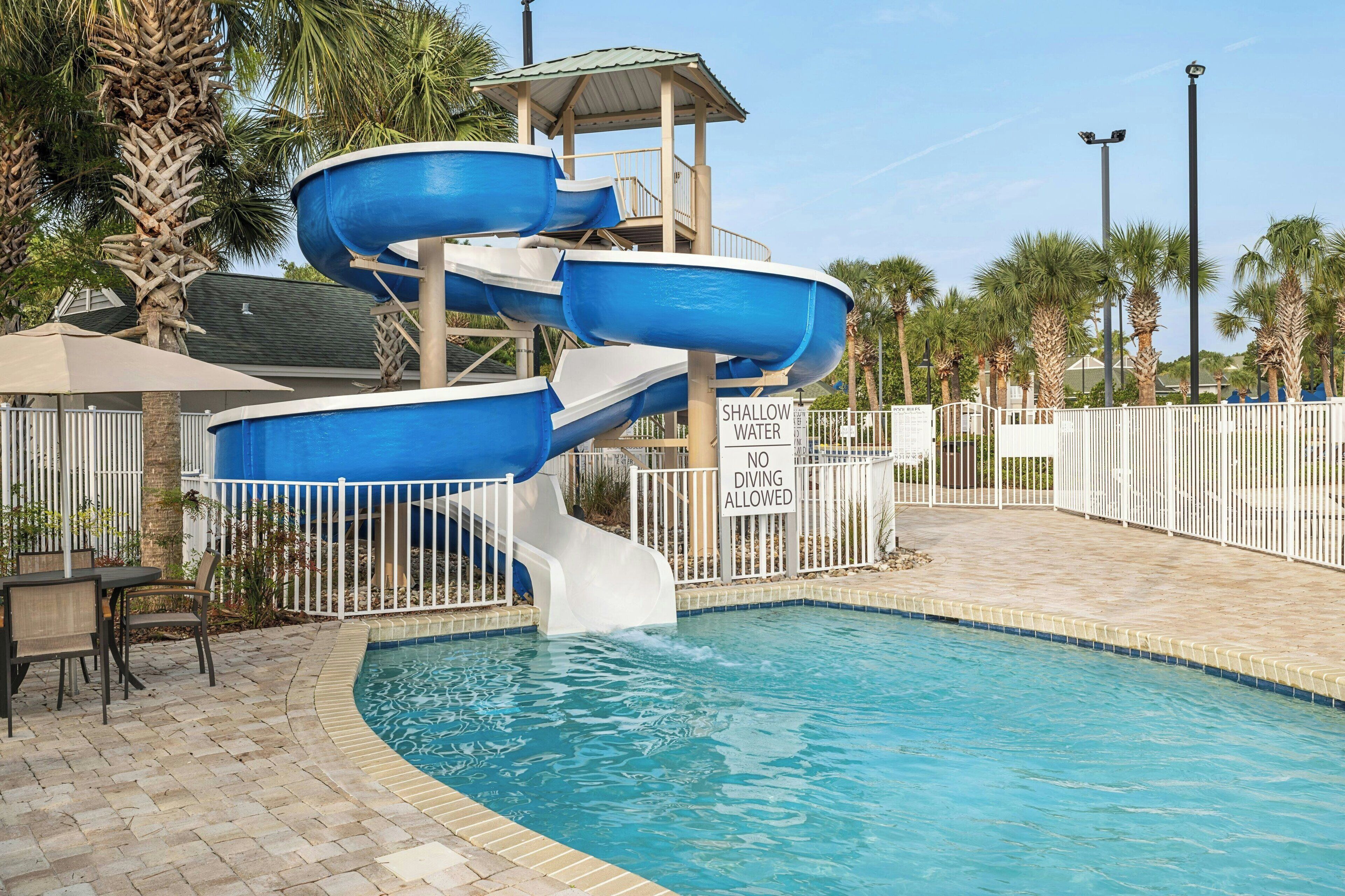 Een waterpark met een blauwe glijbaan in meerdere niveaus en een waarschuwingsbord voor ondiep water.