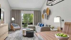 Un salon moderne avec un canapé, un pouf, une table basse et une table à manger sur laquelle se trouve un saladier rempli de pommes. On y trouve également une fenêtre avec des rideaux, une lampe et un luminaire au plafond.