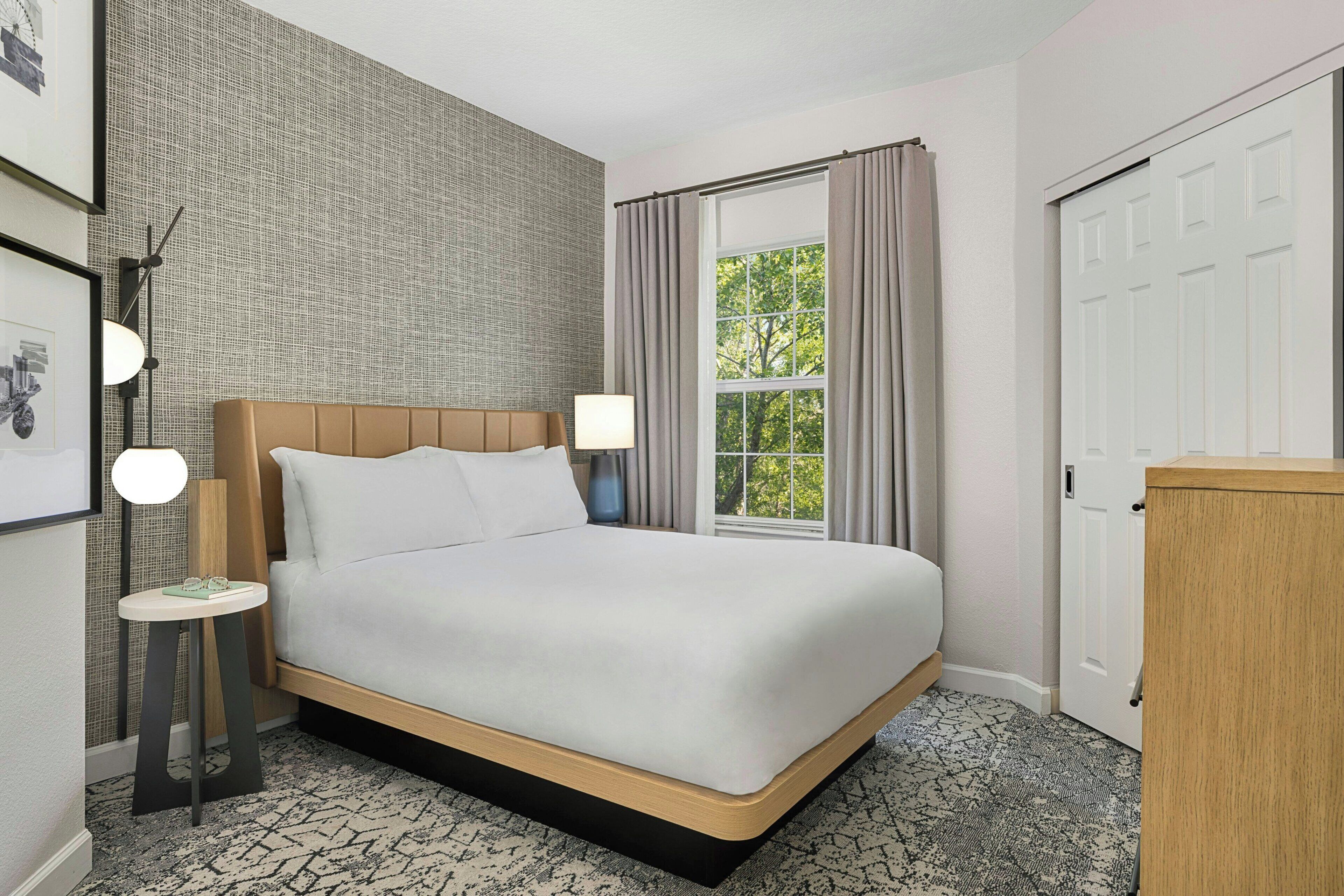 A hotel room with a large bed, a window with curtains, a bedside table with a lamp, and a wall with a textured pattern.