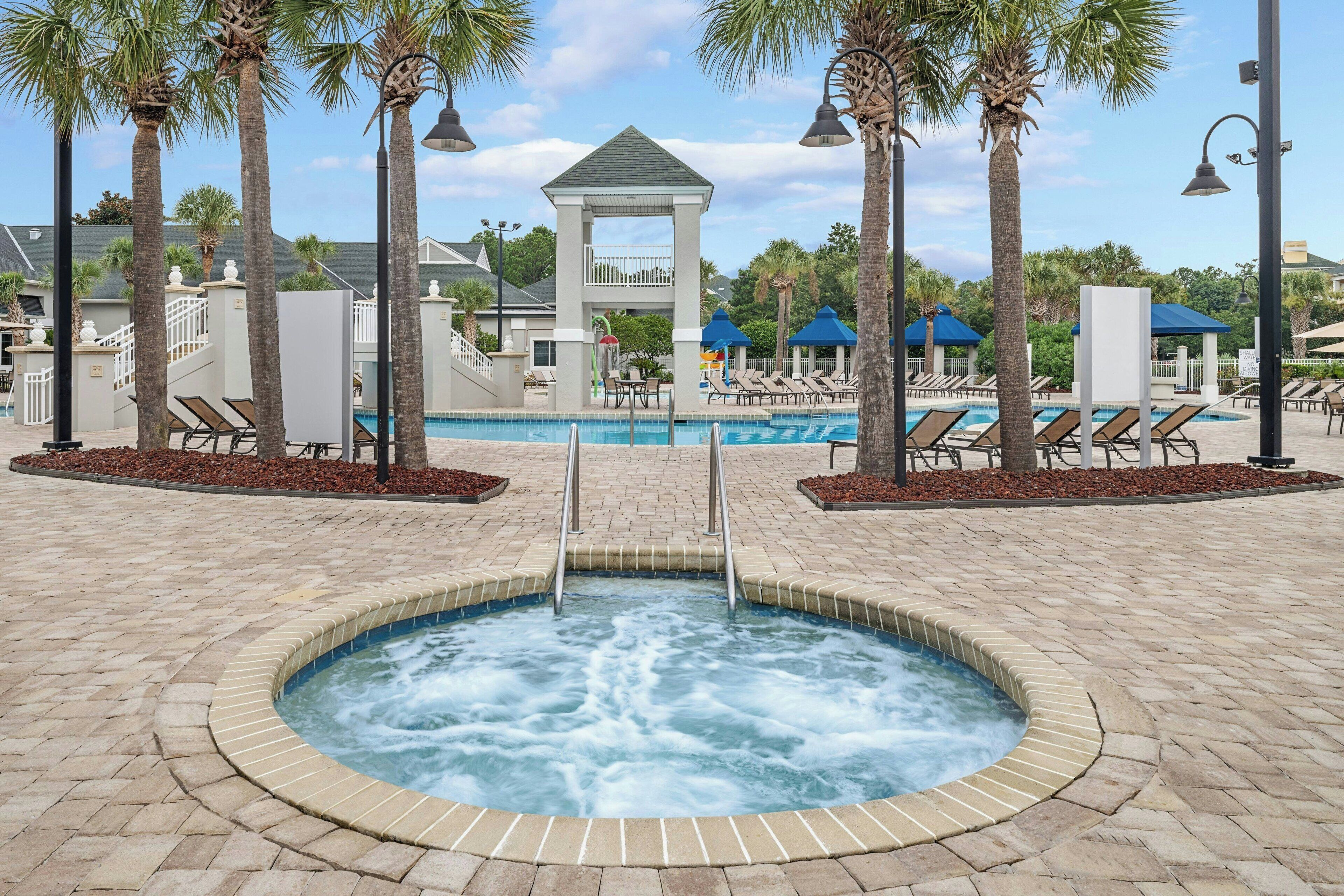 Ein Whirlpool, umgeben von Liegestühlen und Palmen, mit einem Pavillon und einem Pool im Hintergrund.