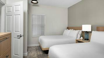 A hotel room with two beds, a bedside table with a lamp, a window with blinds, a door, and a patterned carpet.