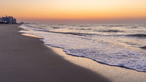 Une plage avec des vagues au coucher du soleil, des bâtiments en arrière-plan et un ciel dégagé.