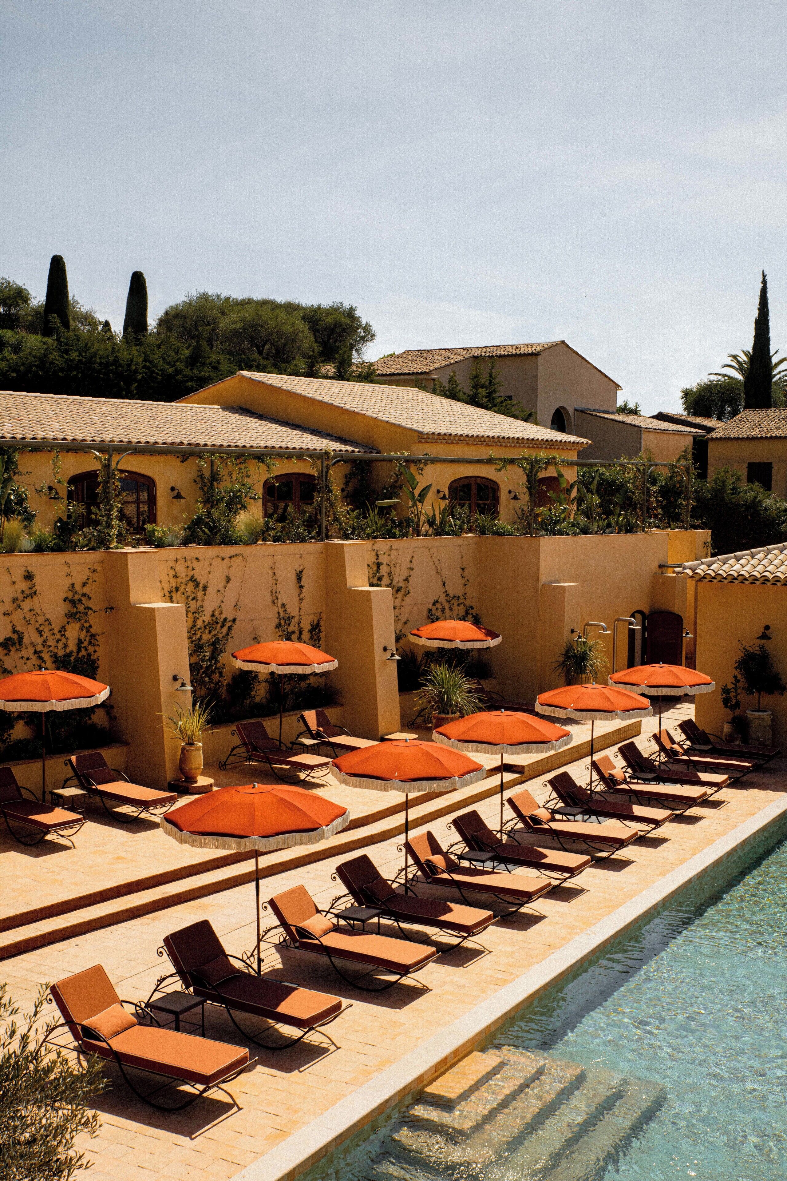 A poolside area with sun loungers and orange umbrellas, surrounded by a building with a tiled roof and greenery.