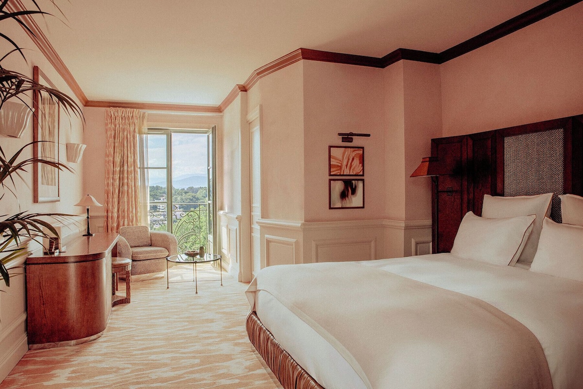 A hotel room with a large bed, a small seating area with a table, a view of the outdoors through a window, and a fireplace.