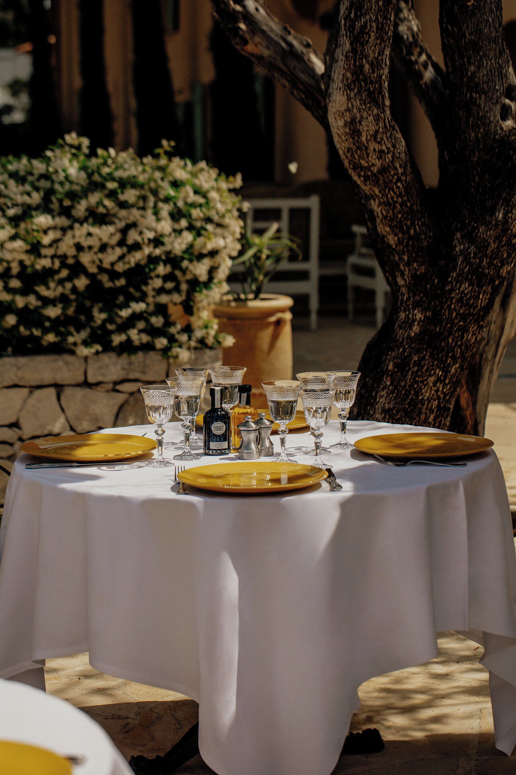 An outdoor dining table set with yellow plates and clear glassware, surrounded by a stone wall and a large tree.