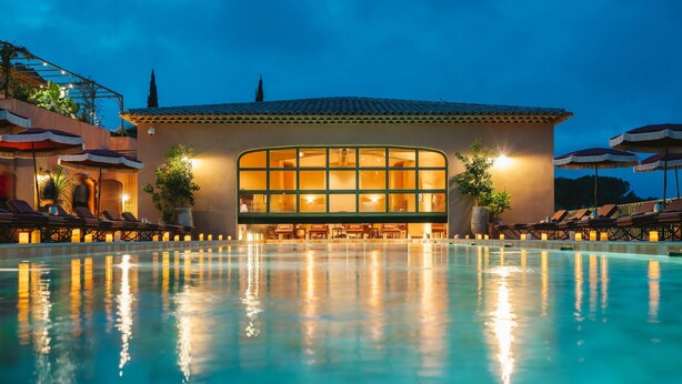 Un hotel con una piscina grande, asientos al aire libre y una fachada iluminada.