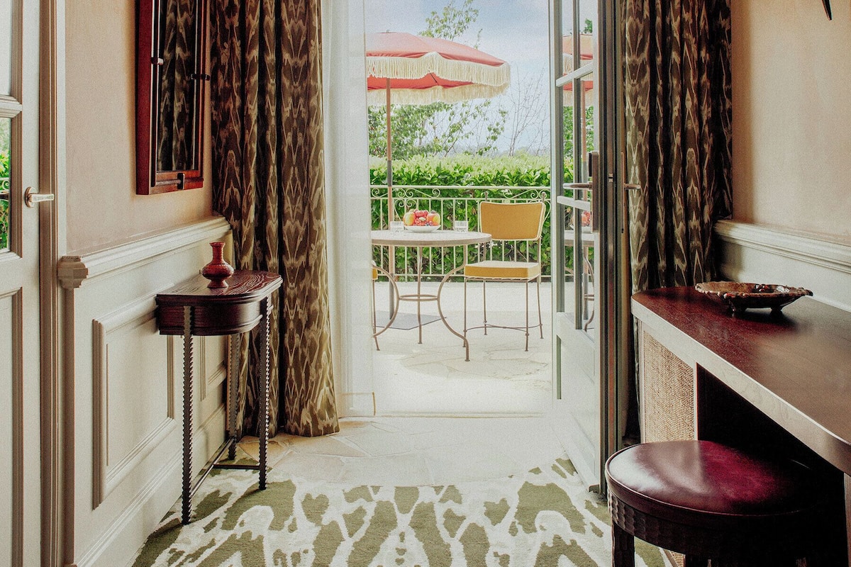 A hotel room with a view of an outdoor patio, a wooden table, a chair, and a red umbrella.