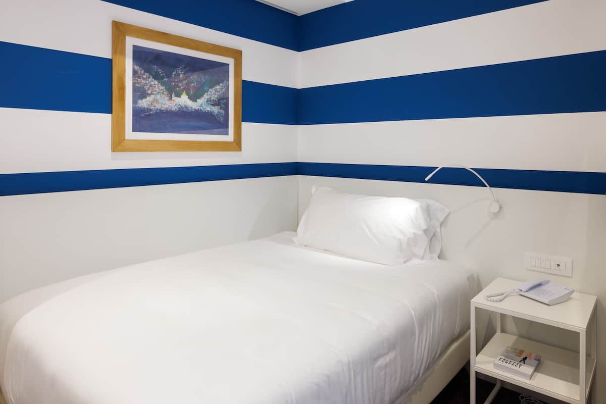 A hotel room with a bed, a bedside table with a phone and a magazine, and a painting on the wall.