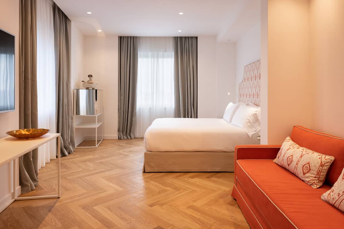A modern hotel room with a large bed, a red sofa, a wooden floor, and a small table with a bowl.