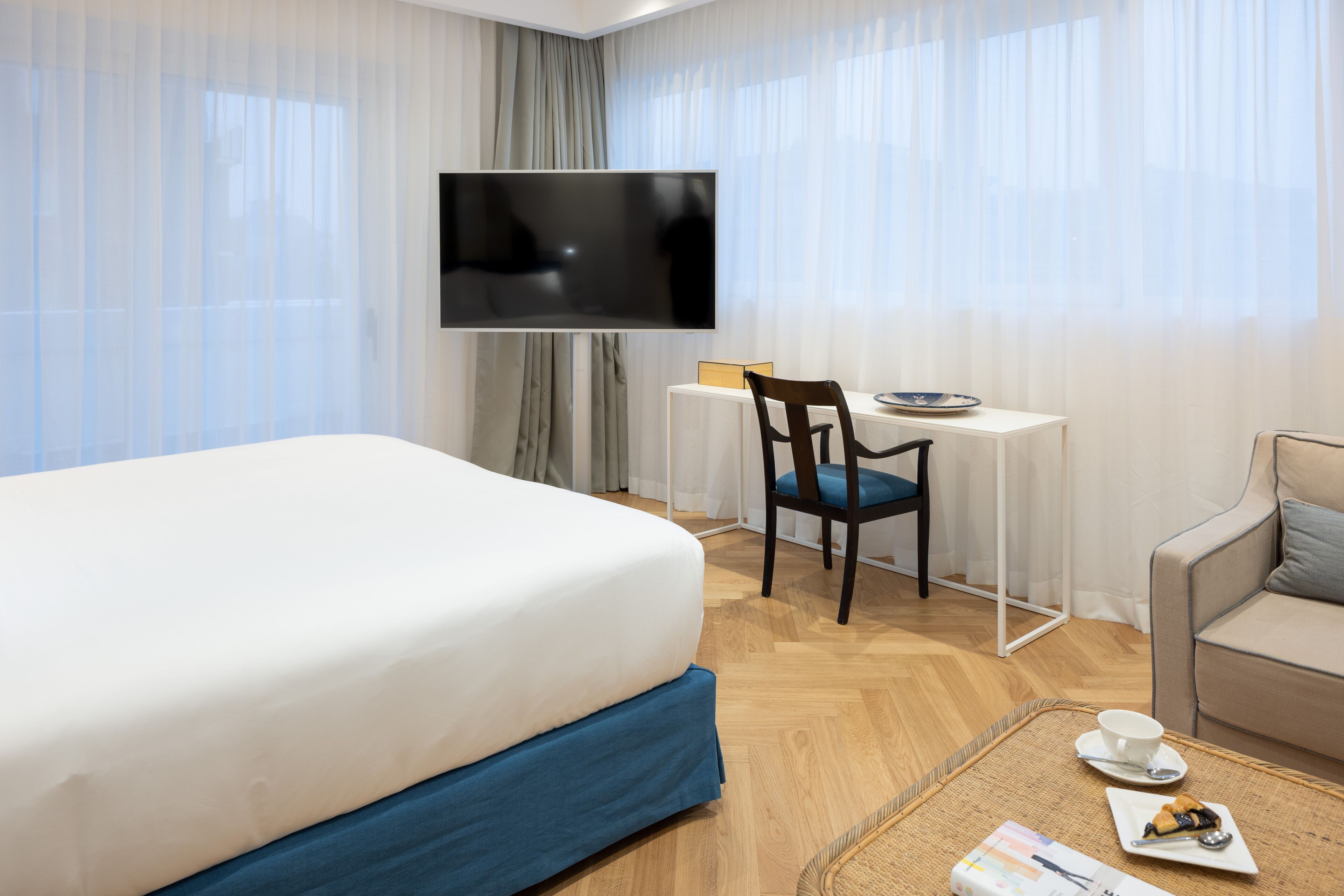 Habitación de hotel con cama, escritorio, silla, televisor, sofá y zona de comedor con taza y plato.