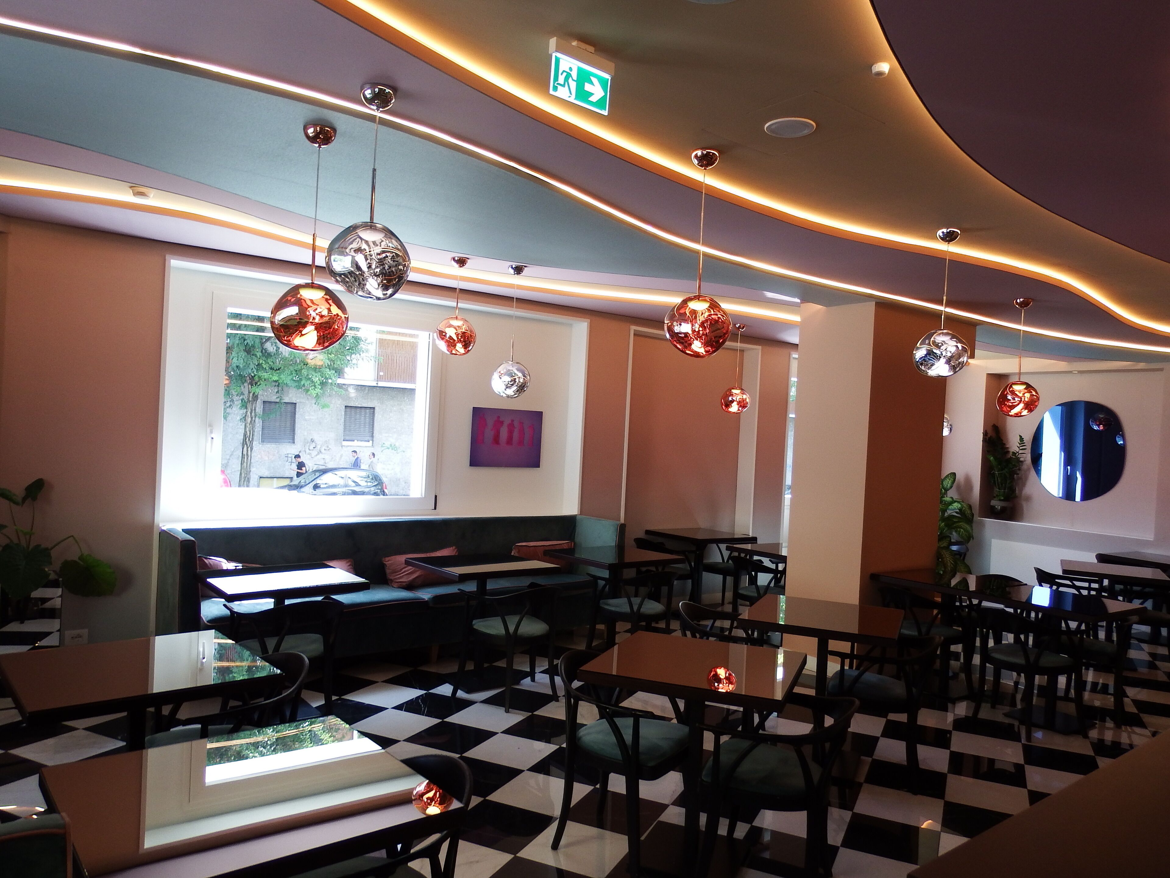 Een restaurant met geruit vloerkleed, meerdere tafels en stoelen, hangende bolvormige lampen en uitzicht op de straat door een raam.