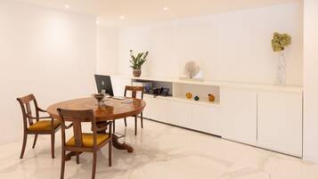 A modern dining area with a round wooden table, two chairs, a laptop, a potted plant, and a shelf with decorative items.