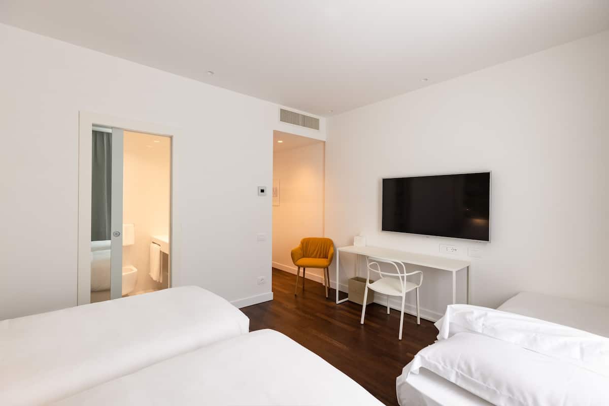 A modern hotel room with two beds, a desk, a TV, and a bathroom visible through an open door.