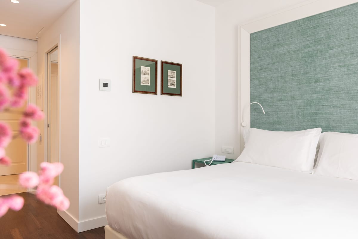 A hotel room with a bed, a green headboard, a nightstand with a phone, and two framed pictures on the wall.