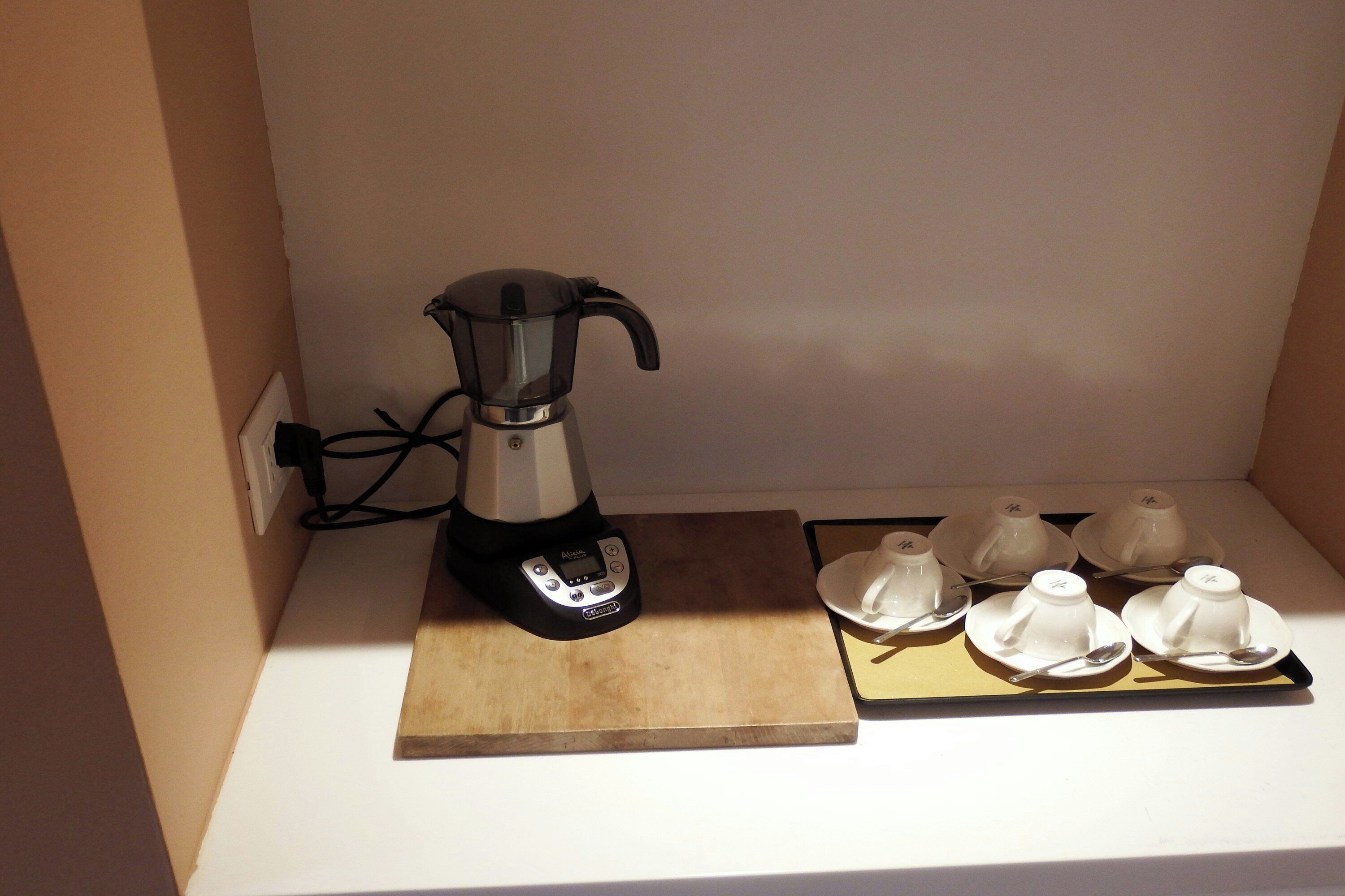 A coffee maker on a wooden board with four white cups and spoons on a tray.