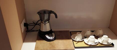 A coffee maker on a wooden board with four white cups and spoons on a tray.