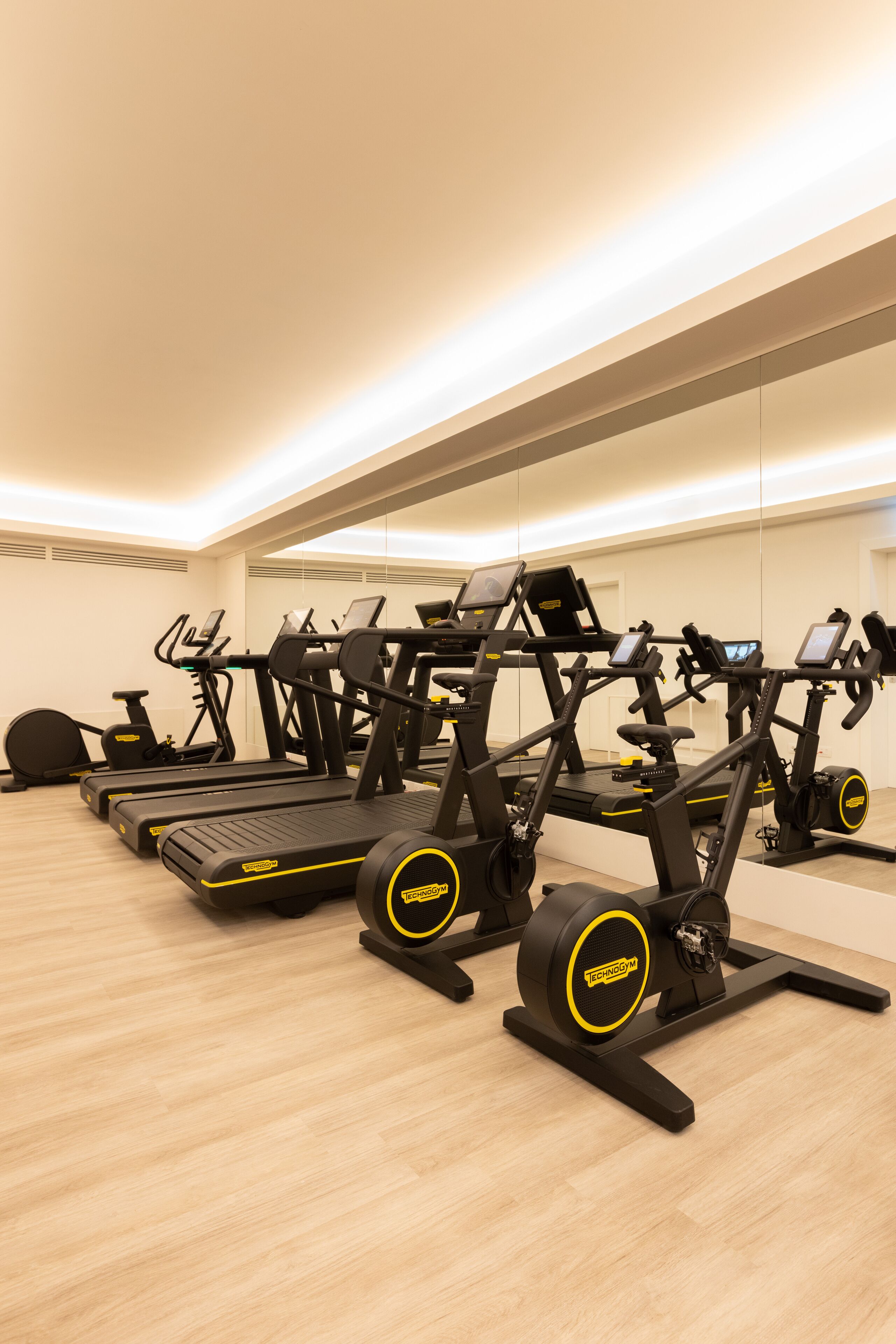 A gym with multiple exercise machines, including treadmills and stationary bikes, arranged in a spacious room with a wooden floor and a large mirror on one wall.