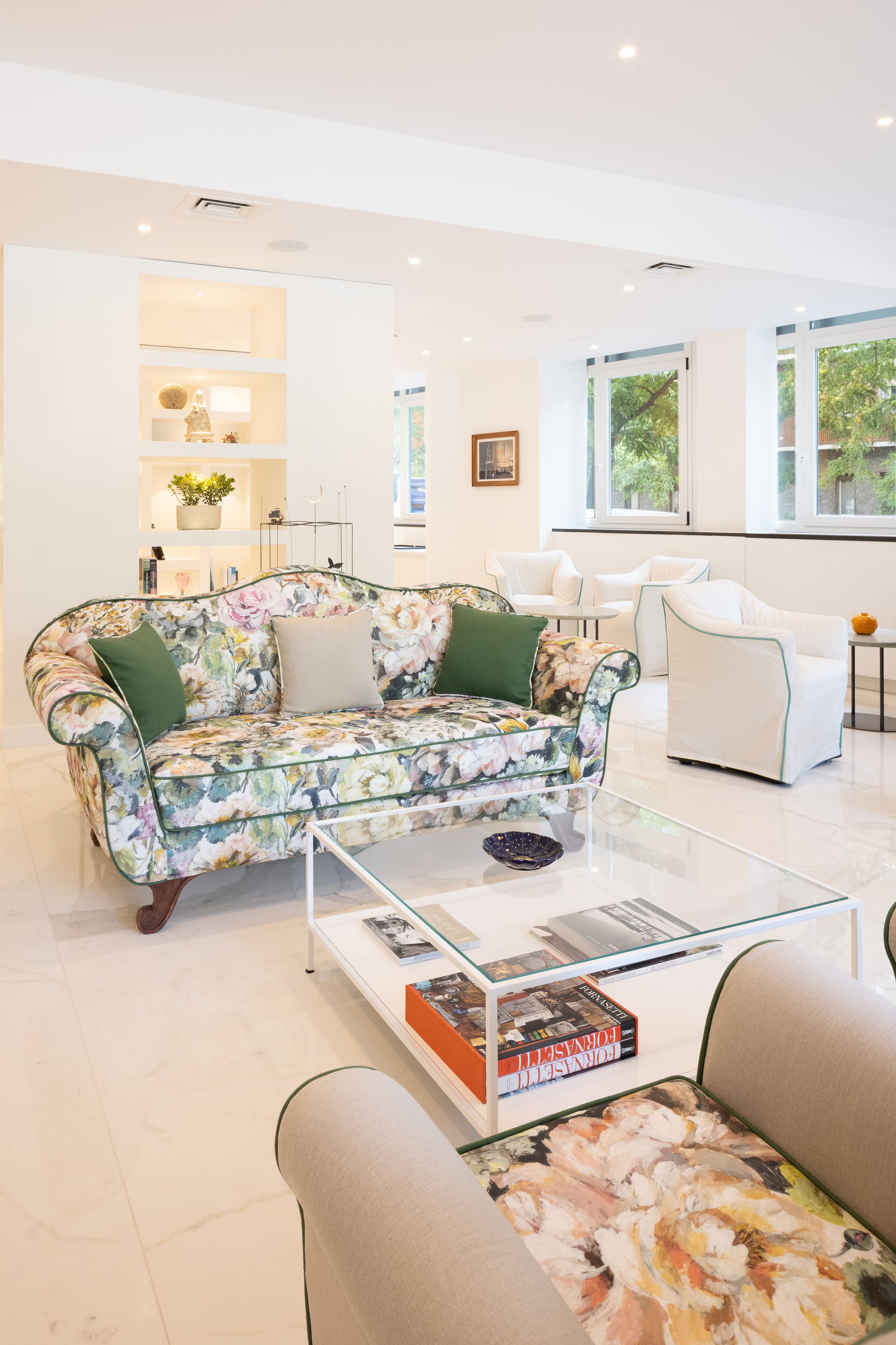 Uma sala de estar moderna com um sofá estampado floral, uma mesa de centro de vidro e uma estante com livros.