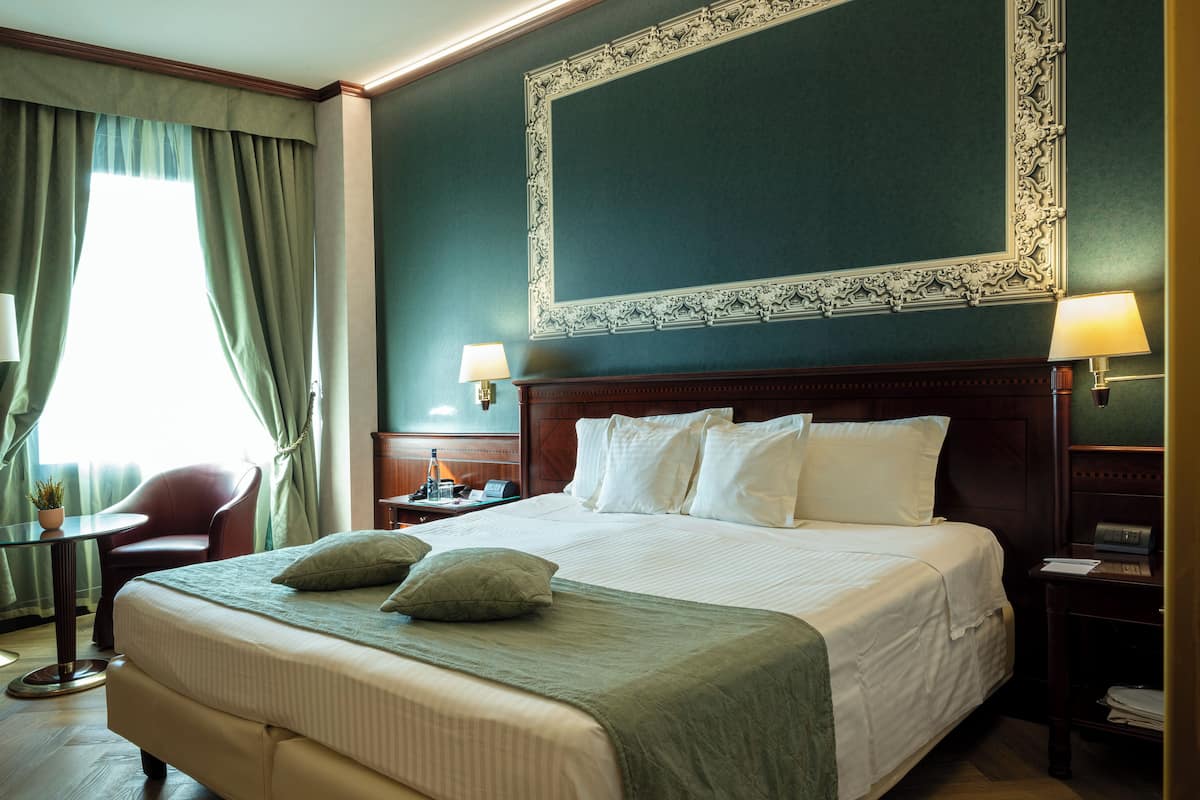 Ein Hotelzimmer mit einem großen Bett, zwei Nachttischlampen, einem Nachttisch, einem Sessel und Blick ins Freie durch ein Fenster mit Vorhängen.
