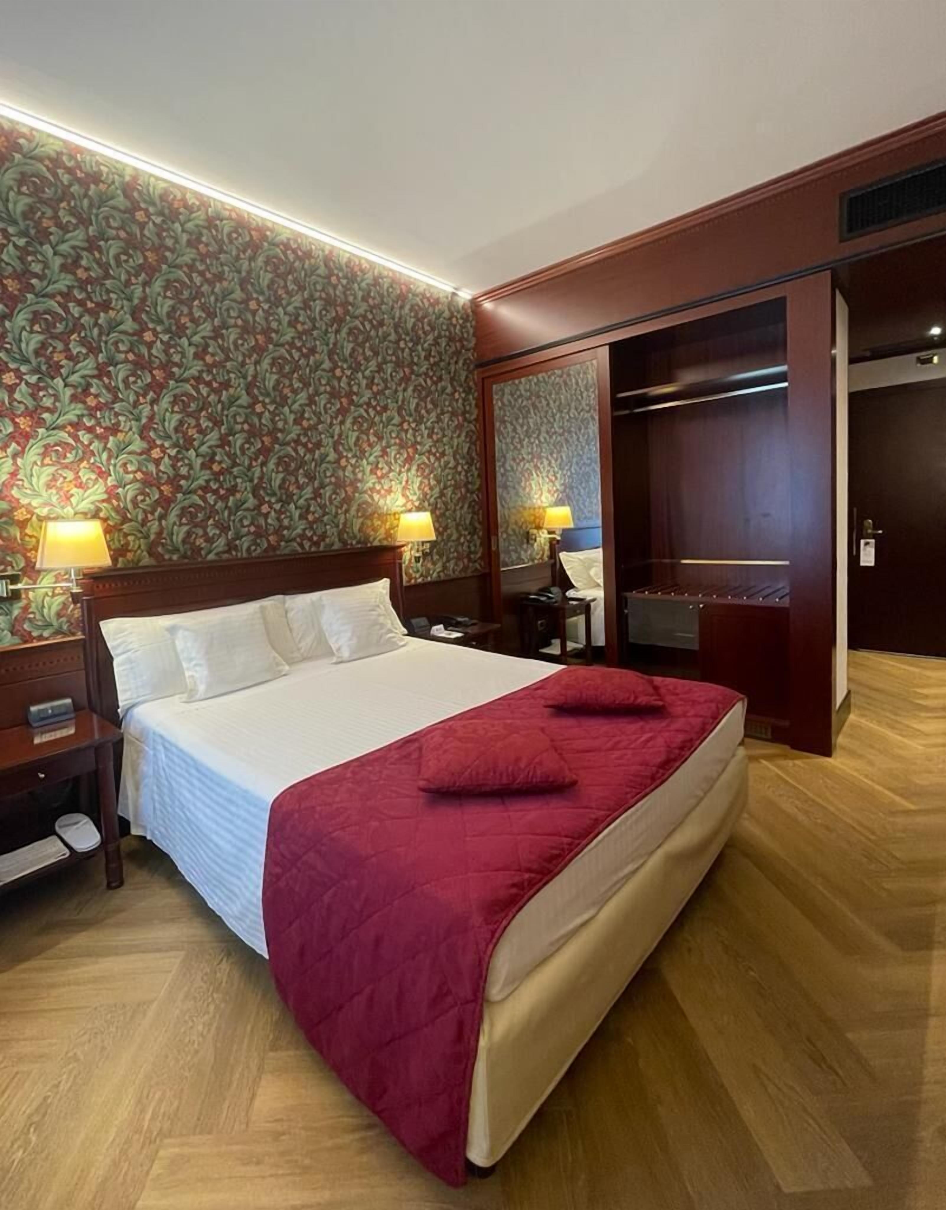 Ein Hotelzimmer mit einem großen Bett, zwei Nachttischlampen, einem hölzernen Kopfteil, einer roten Tagesdecke, einem Holzboden und einem blumigen Wandpapier.