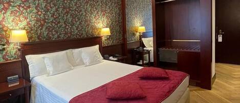 Ein Hotelzimmer mit einem großen Bett, zwei Nachttischlampen, einem hölzernen Kopfteil, einer roten Tagesdecke, einem Holzboden und einem blumigen Wandpapier.