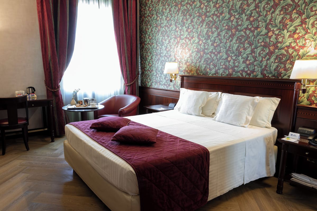Ein Hotelzimmer mit einem großen Bett, einem Schreibtisch mit Stuhl, einem kleinen Tisch mit Lampe und einem Fenster mit Vorhängen.