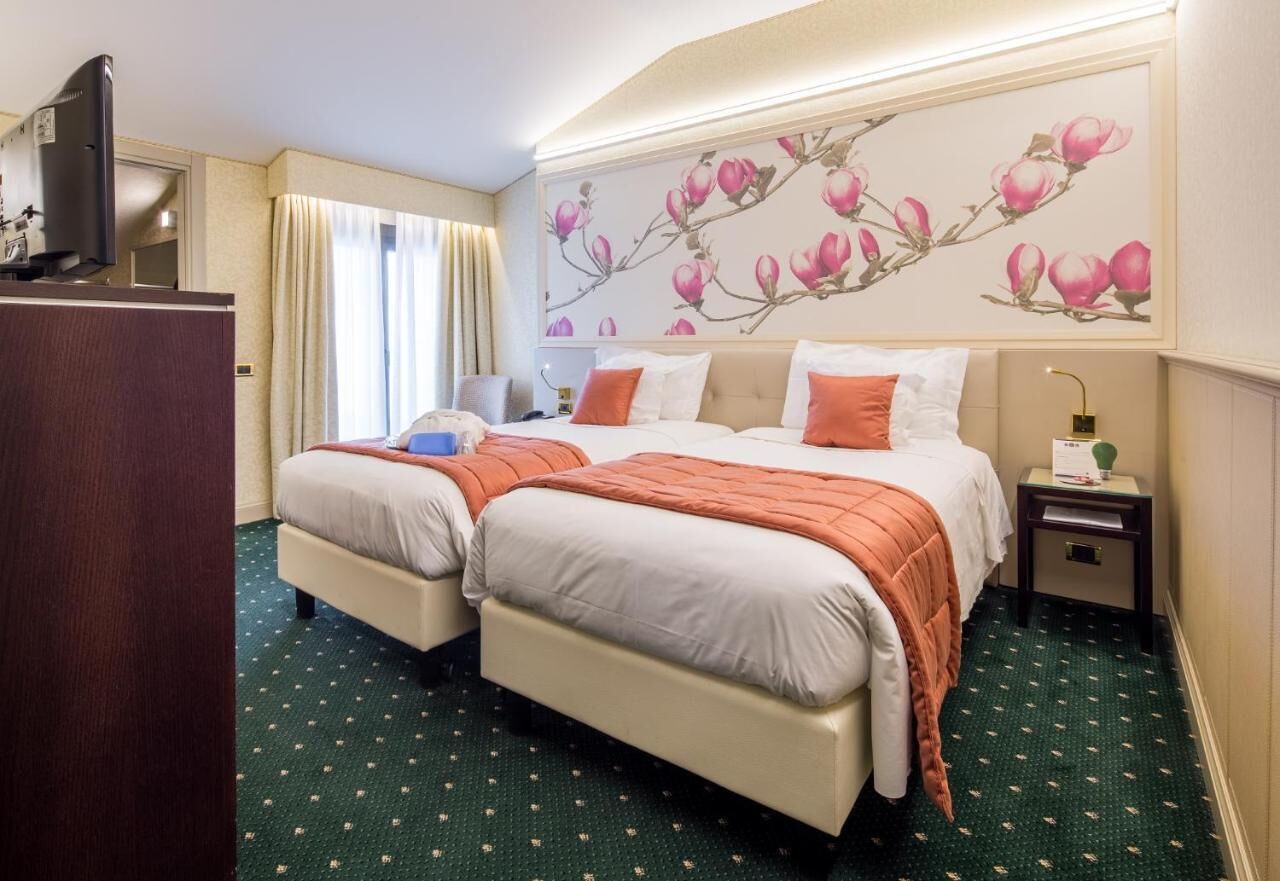 Habitación de hotel con dos camas, un escritorio, un televisor y una pared con un mural floral.
