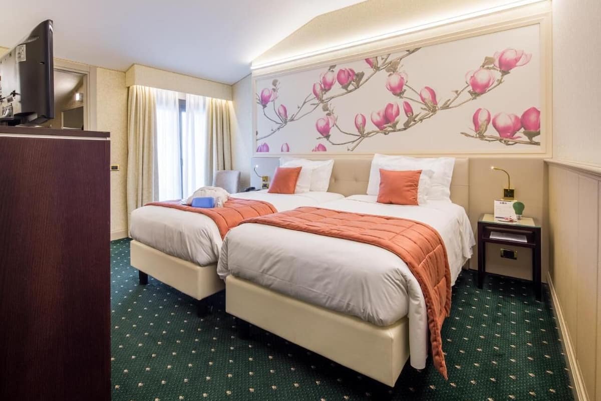Habitación de hotel con dos camas, un escritorio, un televisor y una pared con un mural floral.