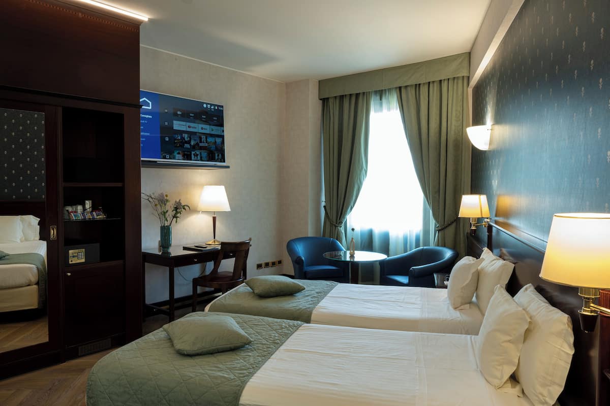 Ein Hotelzimmer mit zwei Betten, einem Schreibtisch, einem Stuhl, einem Fernseher und einem Fenster mit Vorhängen.