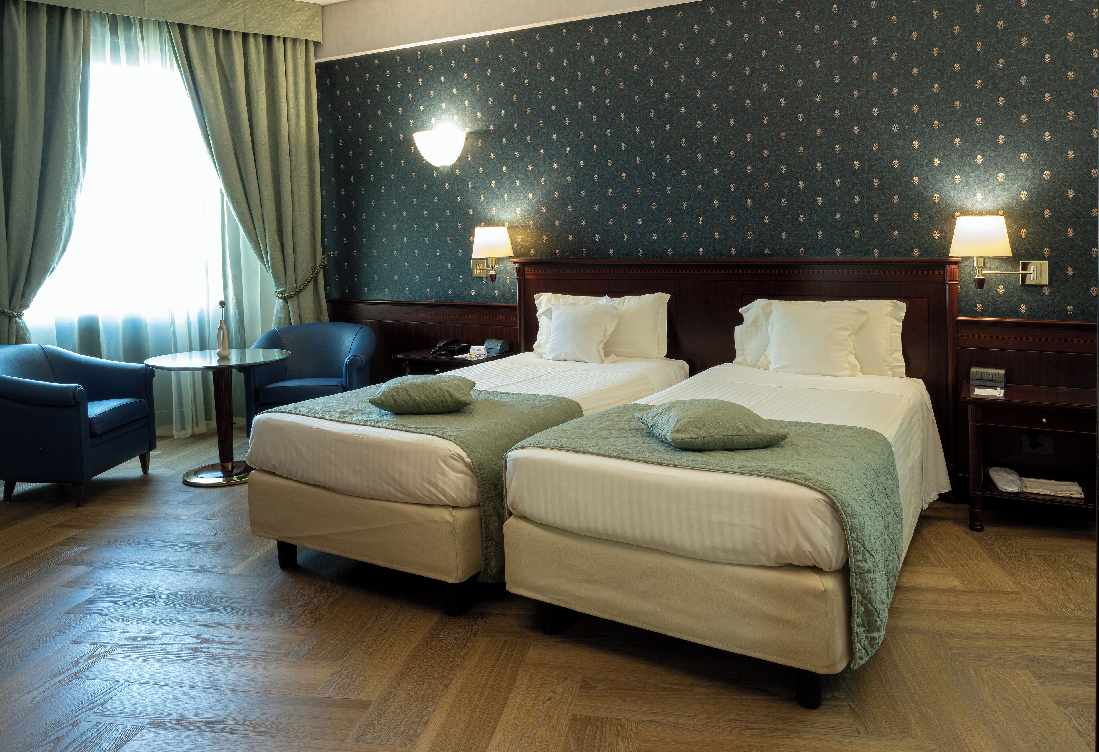 A hotel room with two beds, a sitting area with a chair and a small table, and a large window with curtains.
