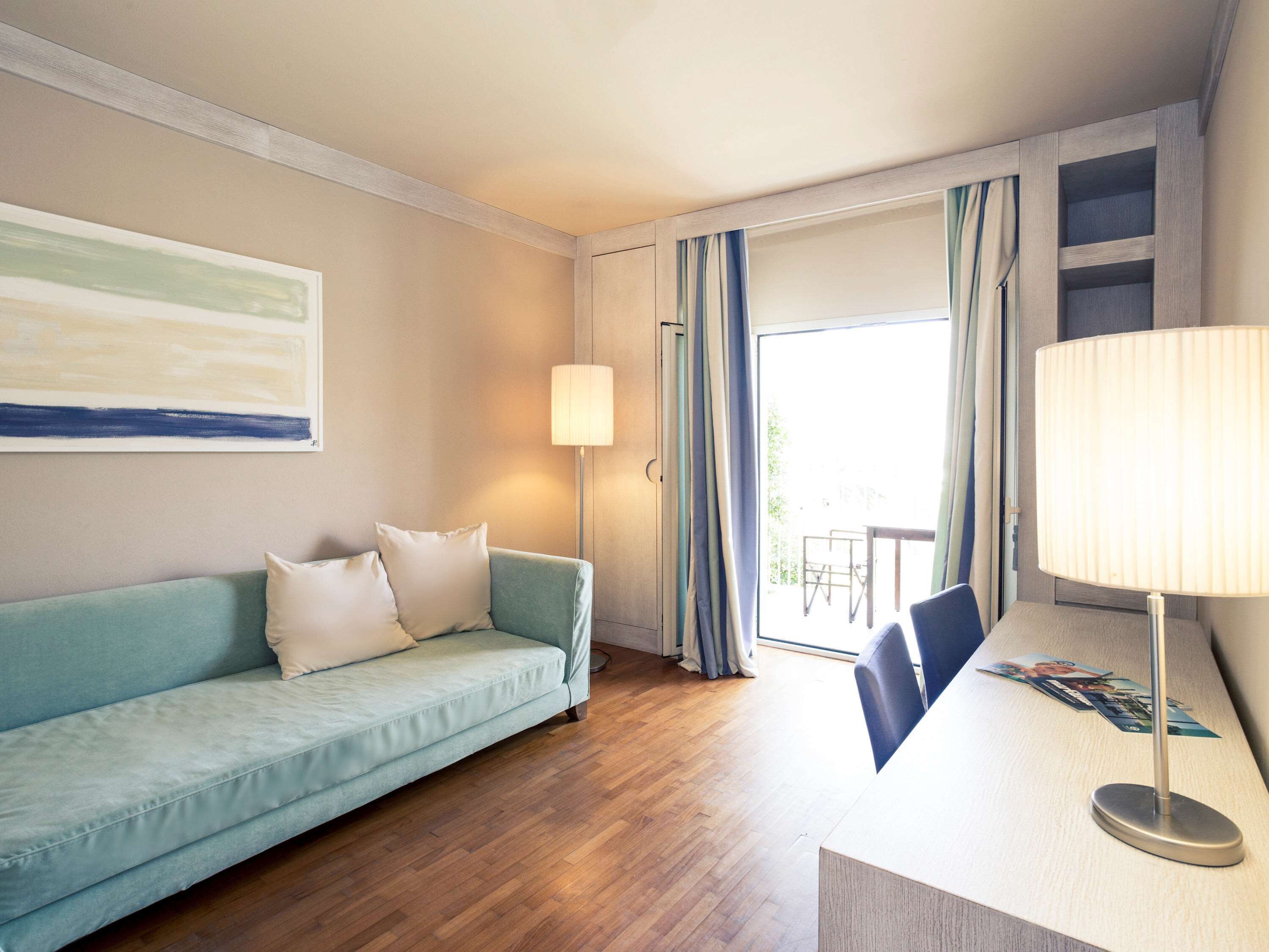 Habitación de hotel con un sofá azul claro, una lámpara de pie, suelo de madera, un ventanal con cortinas y un cuadro en la pared.