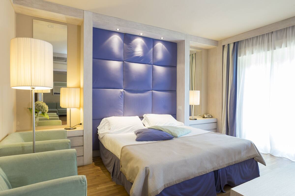 Une chambre d’hôtel moderne dotée d’un grand lit, d’un coin salon avec des fauteuils, d’une table de chevet avec une lampe et d’une grande fenêtre avec des rideaux.